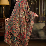 A woman models a large, ornate shawl or scarf with an intricate multicolor paisley and floral pattern in red, green, gold, and brown tones, set against a dark, elegant interior background with chandeliers.