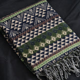 A folded woven throw blanket with geometric patterns in navy, cream, green, and brown, featuring fringed edges, displayed on a dark fabric background.