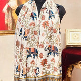 A cream-colored scarf with a colorful print of elephants, flowers, and traditional patterns, displayed on a black mannequin torso in an ornate room setting.
