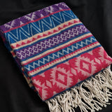 A folded woven throw blanket with a colorful geometric pattern and cream fringe, displayed on a black fabric background.