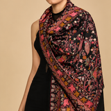 A black embroidered shawl or scarf with intricate floral and bird motifs in pink, orange, and gold, shown on a model in two poses and in a close-up detail panel against a beige studio background.