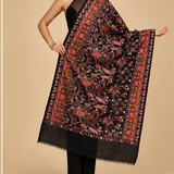 A black embroidered shawl or scarf with intricate floral and bird motifs in pink, orange, and gold, shown on a model in two poses and in a close-up detail panel against a beige studio background.