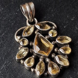 A vintage-style silver pendant featuring a central rectangular yellow gemstone surrounded by smaller marquise and round yellow stones, set against a dark textured background.
