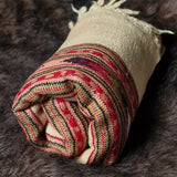 A rolled woven throw blanket with a cream base and a striped multicolor pattern in red, green, black, and beige, resting on a dark brown faux fur surface.