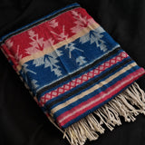 A folded woven throw blanket with fringe, featuring a Southwestern-style pattern in red, blue, cream, and black on a dark background.