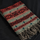A woven Turkish-style throw blanket with geometric patterns in red, dark green, and beige, featuring fringed tassels on one end, laid flat on a black background.