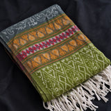 A folded woven textile throw blanket with fringe, featuring horizontal bands of gray, orange, red, and olive green with geometric and floral patterns, laid on a dark fabric background.
