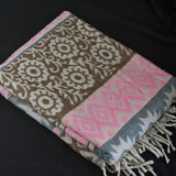A folded woven throw blanket with a brown and cream floral pattern, a pink and cream geometric diamond border, and cream fringe tassels, displayed on a black background.