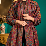 A model is shown wearing a long, open-front shawl or lightweight coat with an intricate multicolor paisley and floral pattern in deep red, blue, green, and gold tones, set against a dark teal background with ornate gold furniture.