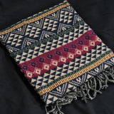 A folded woven throw blanket with geometric patterns in navy, cream, burgundy, and olive green, featuring fringed edges, laid flat on a dark background.