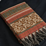 A folded woven throw blanket with geometric patterns in brown, beige, coral red, and teal, featuring fringed tassels, displayed on a dark background.