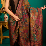 A model wearing a large, ornate shawl with intricate paisley and floral patterns in deep red, gold, teal, and brown, styled over a black dress in an opulent room with a green wall and chandeliers.