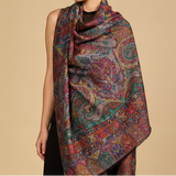 A model is shown in three views wearing a long, lightweight shawl with an intricate multicolor paisley pattern. The shawl features a dense mix of colors including red, blue, green, and gold on a dark base, and is styled over a black outfit against a solid beige studio background.