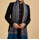 A four-panel collage showing a model wearing a large patterned shawl scarf in different draped poses. The scarf has a deep blue base with intricate multicolor paisley and floral motifs in red, purple, and gold, finished with a solid blue border.