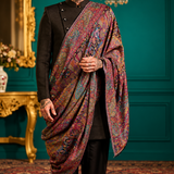 A man wearing a black traditional outfit and a vibrant, multicolored patterned shawl draped over his shoulder, standing in an ornate room with teal walls, gold furniture, and a chandelier.