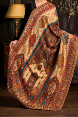 A woman models a heavily patterned beige and brown sari with intricate red, blue, and gold motifs, posed in an opulent room with chandeliers and a lamp.