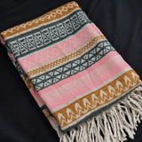 A folded woven throw blanket with a Southwestern geometric pattern in pink, mustard yellow, dark gray, and white, featuring cream fringe tassels, displayed on a black background.