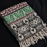 A folded woven throw blanket with cream, black, green, and burgundy patterns and fringe tassels, displayed on a black fabric background.