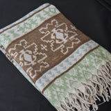 A folded woven throw blanket with a Southwestern geometric pattern in brown, sage green, cream, and light blue, featuring a fringed edge, displayed on a dark background.