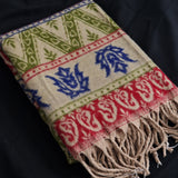 A close-up of a woven textile with fringe, featuring cream, green, red, and blue geometric and floral patterns on a dark fabric background.