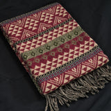 A folded woven throw blanket with geometric tribal patterns in burgundy, beige, and black, featuring gray fringe tassels, laid flat on a dark background.