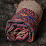 A rolled woven textile, likely a blanket or throw, made from natural tan burlap with a vibrant striped pattern in pink, blue, orange, and green, displayed on a dark textured fur surface.