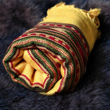 A rolled yellow woven textile with fringed edges and a striped pattern in green, red, and dark brown, displayed on a dark textured faux fur surface.