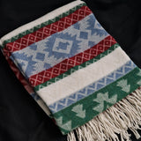 A folded woven throw blanket with a Southwestern geometric pattern in red, blue, green, and cream, featuring fringe on one end, laid on a black background.