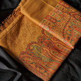 A close-up of a patterned shawl or scarf in warm golden yellow, red, orange, and teal paisley and floral motifs, laid over a black fabric background.