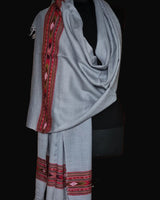 A light gray shawl with red, pink, and black geometric embroidered borders, draped on a black dress form against a wooden door.
