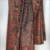 A heavily patterned shawl or scarf with intricate paisley and floral motifs in red, orange, yellow, and blue on a dark background, hanging against a plain white wall.