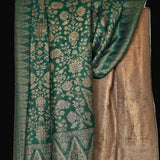 A green and gold Banarasi silk saree with intricate floral and geometric patterns, draped against a solid black background.