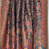 A draped silk scarf with an intricate, dense traditional pattern in black, red, orange, and gold, featuring peacock motifs and detailed figures, hanging from a white rod against a white wall.