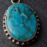 A round turquoise cabochon pendant with a silver beaded border, shown close-up on a dark textured background.