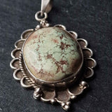 A silver-toned scalloped pendant necklace featuring a round green and brown marbled stone cabochon, photographed close-up against a dark textured background.