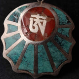 A close-up of a decorative metal pendant or medallion with turquoise inlay and a central red circular area featuring a white symbol, photographed against a dark background.