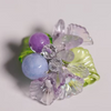 A small floral embellishment or brooch with translucent lavender petals, a light blue bead, a purple bead, and green leaf accents on a plain light background.