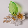 A decorative floral brooch featuring a translucent green leaf, white flower petals, and faceted amber and clear crystal drops on a silver-toned base, photographed on a plain light background.