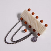 An off-white carved bone or horn pendant with carved diagonal lines, small circular details, and multiple small orange-brown beads, attached to a dark silver chain with a round medallion charm, on a light gray background.