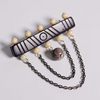 A Viking-style beaded brooch with a rectangular bar engraved with geometric patterns, adorned with cream-colored beads and two hanging chains ending in round beads, shown on a light gray background.