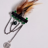 A decorative feather hair accessory with brown and cream feathers, a black bar, green beads, silver chains, and a round silver coin pendant, photographed on a plain light background.