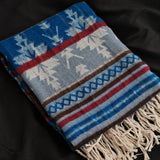 A folded woven throw blanket with cream fringe, featuring a Nordic-inspired pattern of snowflakes, trees, and stripes in blue, gray, red, brown, and cream on a black fabric background.