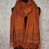 A rust-orange woven scarf with fringed ends and a striped patterned border, draped on a black mannequin torso against a textured gray background.