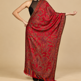 A woman modeling a red shawl with intricate gold and green paisley patterns, draped over her shoulders against a solid beige background.