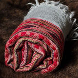A rolled woven textile throw or blanket with fringed ends, featuring a striped pattern in red, pink, beige, and brown with a light gray section, displayed on a dark brown furry surface.
