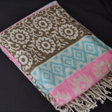 A folded patterned throw blanket with cream, brown, light blue, and pink woven designs and fringed edges, displayed on a dark background.