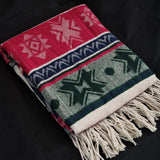 A folded woven throw blanket with a Southwestern geometric pattern in red, navy, green, and cream, featuring white fringe tassels, displayed on a black fabric background.