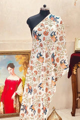 A draped saree displayed on a black mannequin in an ornate indoor setting. The saree has a cream base with a dense multicolor floral and paisley print in orange, blue, and green.
