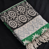 A folded woven throw blanket with cream floral patterns on a black section, a light gray diamond pattern on a cream section, and bright green borders, shown on a black fabric background.