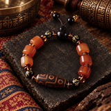A beaded bracelet displayed on a dark stone slab, featuring translucent orange-red agate-like beads, a brown patterned cylindrical bead, black beads, and small gold-toned spacer beads. The bracelet is styled with traditional decorative props including a brass bowl and patterned textile.